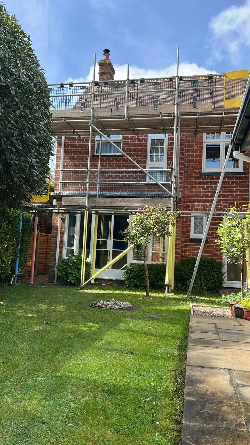 Rear-elevation domestic scaffold over a garden with conservatory, hi-vis edge protection on the eaves lift